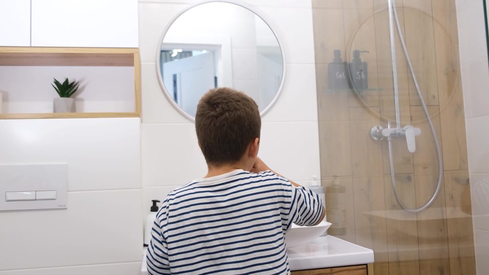 Child washing up at a bathroom sink during a back-to-school morning routine, with a glass-enclosed shower nearby